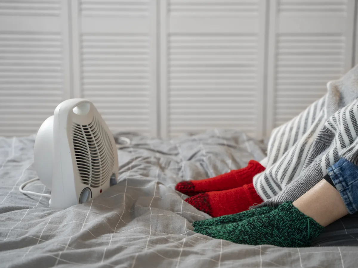 Feet in knit socks under a blanket on a bed, warming up in front of a small space heater